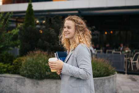 Young woman student dressed in casual clothing walking through the city, drinking coffee to go and enjoying free time outdoors. Smart device.の写真素材