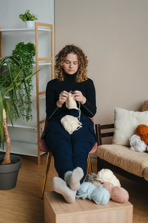 Young concentrated curly-haired woman holding needles sitting on armchair and enjoying knitting....