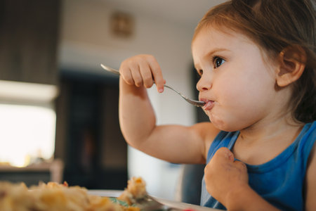 Adorable girl eating meal with fork at home sitting on high-chair, indoors. Childhood growing up and development concept.の写真素材