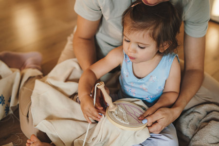 Small baby girl playing while her mother embroidering, doing job or hobby sitting on the floor in living room. Family, candid domestic life with baby. Authentic lifestyleの写真素材