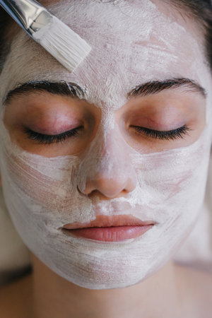 Crop young female with closed eyes applying facial mask on face with brush