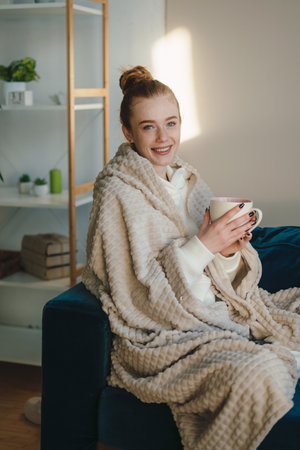 Positive young female with mug of hot beverage smiling and looking at camera while resting in cozy room at homeの写真素材