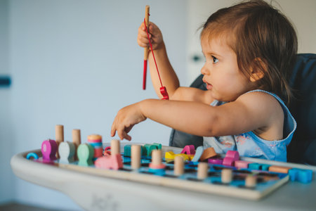 Side view of cute little girl in casual clothes playing with colorful toys while sitting at table in light roomの写真素材