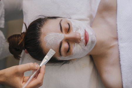 Top view of crop anonymous female applying facial mask on face while lying on bed with closed eyesの写真素材