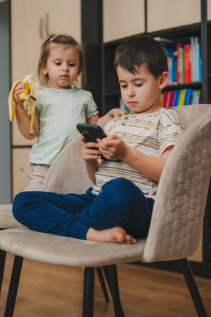 Cute little boy in casual clothes browsing smartphone while sitting at table in cozy living room at home while her sister is eating a banana.の写真素材