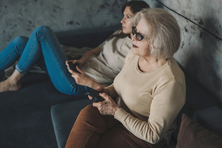 Woman with her grandmother playing video game using controller or joystick while sitting on sofa. Games on console. They plays with wireless controllers.の写真素材