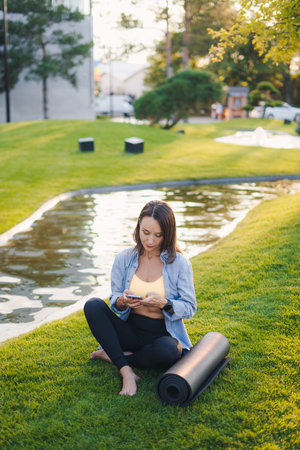Adult woman using smartphone online internet technology after training fitness in summer park. Tourist relax after exercising sport outdoors.の写真素材