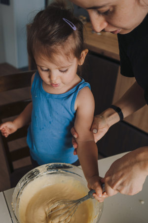 Cheerful mother teaching cute daughter to mix batter with wire whisk in bowl in kitchen. Happy, unaltered, family, togetherness, childhood, food, preparation, learning, lifestyle, home.の写真素材