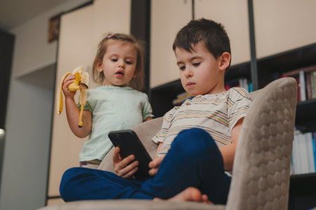 Two little gen Z kids using mobile phone, playing video game for children, relaxing on comfortable armchairs. Digital technology.の写真素材