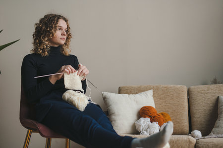 Young creative woman knitting accessories, enjoying hobby work using ribbon yarn. Smiling female creating exclusive handmade decorative details relaxing on armchair at cozy home.の写真素材