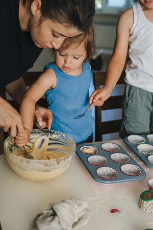 Mother teaching little daughter carefully pouring raw dough for baking muffins in kitchen at home. Homemade cooking.の写真素材