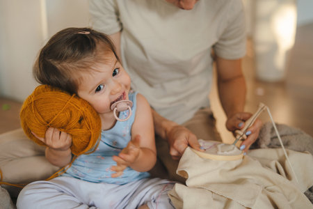 Close up smiling little daughter knitting together with her mother, sitting on floor at home, holding needles. Enjoying leisure time, engaged in creative activityの写真素材