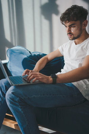 Handsome caucasian man working on laptop computer while sitting on sofa in cozy living room. Freelancer, working from home. Browsing Internet. Using social networks. Having fun.の写真素材
