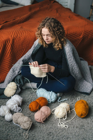 Young curly-haired woman knitting at her leisure time while sitting on the floor in the evening near bed.Cozy home and anti stress hobby.の写真素材