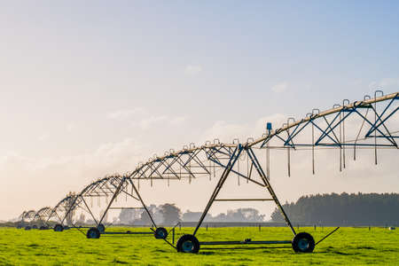 Agriculture modern irrigation. watering spray machine set on the green grassland field with blue sky and warm light scene.の写真素材