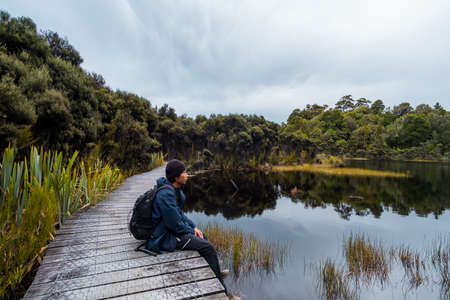 A young Asian man sitting on the path beside a beautiful lake in a cloudy day.