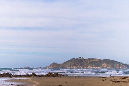 A scene of a beach on a cloudy day with mountain and road to the lighthouse, Nugget point, the...