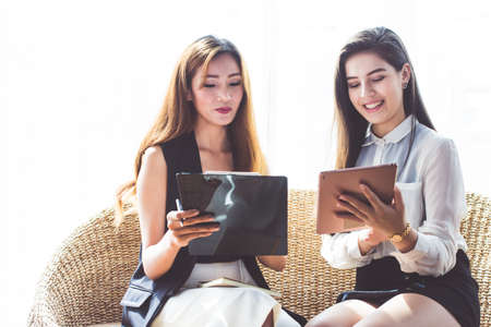 Two Asia Business woman sitting on wooden sofa are smiling, meeting point to discuss, plan and On line Trading of economic cosmetics and work on tabletの写真素材