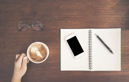 Woman's hand holding cup of coffee with notebook and pen on wooden table. Copy space for text