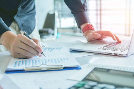 Close up. Hand business holding pen  point to discuss, plan and chart the rise and fall of the economy with laptop on desk,sunset background,Selective focusの写真素材