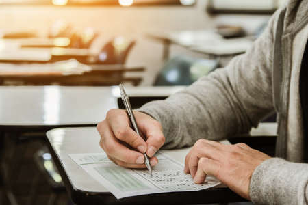 Student holding pencil writing on paper answer sheet.sitting on chair doing final exam attending in examination on classroom.の写真素材