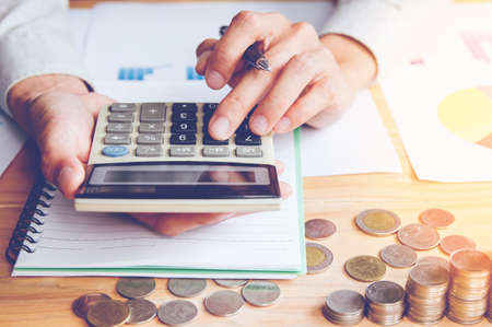 Businessman in grey shirt Hand holding ,Calculation with calculator and Financial data analyzing and counting on calculator in office on wood desk with light sunset background.の写真素材