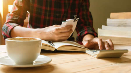 Close up Hand of Asian man in a plaid shirt reading tax, transaction and Calculating numbers using the calculator on table .vintage style.の写真素材
