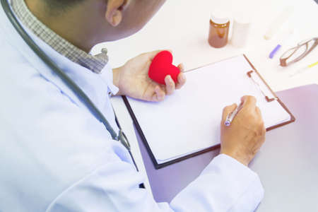 Doctor holding red heart and pen writing report  Treatment Plan on desk with sunset light.Selective focusの写真素材
