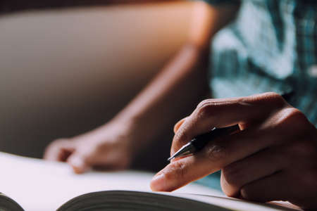 Open book . Man in green plaid shirt holding pen and reading book on table