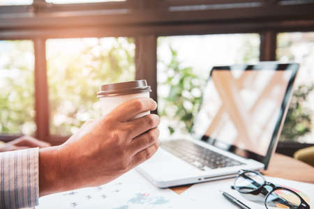 Businessman hands holding cup of  coffee  and using laptop with blank screen on desk in cafe.