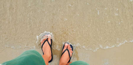 Close up of man in Black slippers feet standing at the beach, with a wave of foaming gentle beneath...