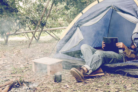 Asian young man sitting is reading a book in outside the tent. Alone camping in forest.