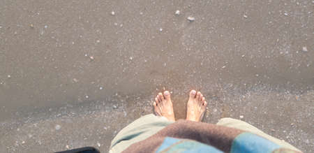 Close up of man in empty feet standing at the beach with a wave of foaming gentle beneath them.Top...