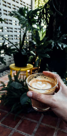 Young man in blue jeans and wearing black shoes sitting on chair holding cup of coffee latte art as...