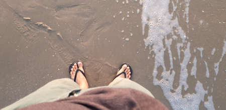 Close up of man in Black slippers feet standing at the beach, with a wave of foaming gentle beneath...