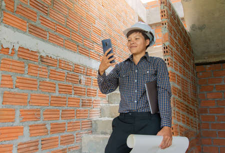 Asian managers of engineer architects are smiling wear white helmet safety on construction site standing check plan interior design and using cell mobile phone in hand-holding blueprint the projectの写真素材
