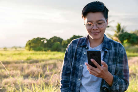 Soft focus,Smiling Asian man wearing plaid shirts and eyeglasses are using a smartphone. Leave screen white space for text and outdoor in the evening and sunset. Young people working on mobile devicesの写真素材