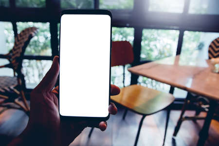 Human left hand using smartphone mobile blank space for your text of white screen display in the cafe coffee shop and wooden table with window mirror vintage interior a light effectの写真素材