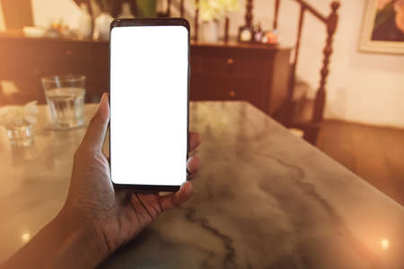 Hand holding mobile phone, blank white screen cell phone on marble table at coffee shop. Mockup...