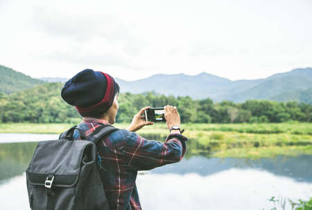 Standing back the Asian man using Camera smartphone Selfie taken of yourself connected to digital camera shooting photo Mountain river landscape. Traveling, Hiking concept.の写真素材