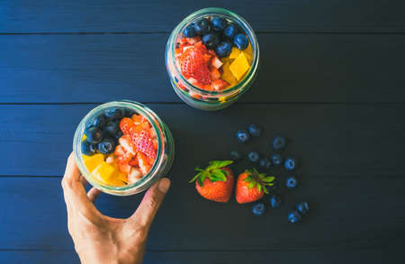 Hand holding overnight oats with fresh mango blueberries and strawberries and in a glass jar on wood table. Breakfast or healthy food,Top view photoの写真素材