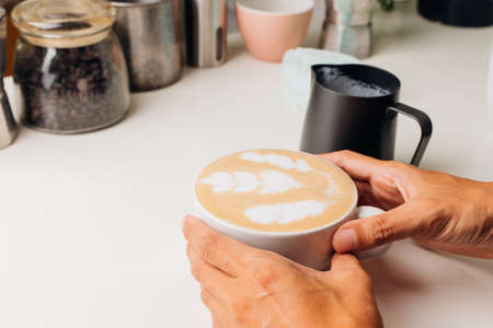 Barista's hand There are hot coffee mugs, latte art and pitcher made of milk froth, and there are coffee-making accessories on the white table.の写真素材
