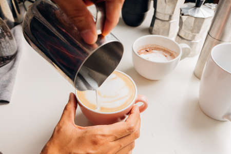 Barista's hand making hot coffee mugs, latte art and pitcher made of milk froth, and there are coffee-making accessories on the white table.の写真素材