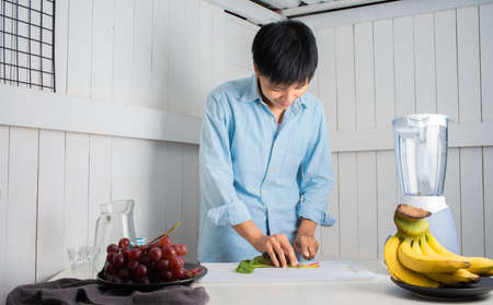 Enjoying Asian man slicing vegetables preparing to Make a Smoothie with Swiss Chard, banana, and red grape at a white kitchen home. people are taking care of themselves.の写真素材