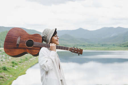A man wearing a hat and casual carrying a guitar on his shoulder is smiling and laughing by the calm river. People, music, learning, and nature outside. music therapyの写真素材