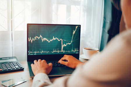 Happyness Asian young man 30s looking stock tickers or graphs cryptocurrency trading platforms on laptop with a cup of coffee, calculator, notebook on the table at home office in the morning.の写真素材