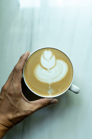 Left hand holds a cup of hot latte art coffee. Homemade coffee, placed on marble vertical photo