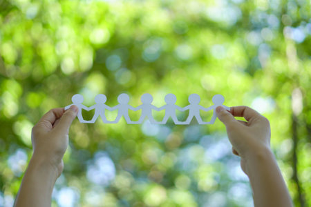 Team of connected paper cut people held by hands, symbolizing teamwork unity and collaboration, against a vibrant green outdoor background representing business support community.の写真素材