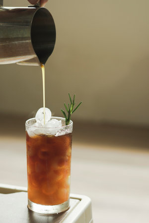 hand hold pitcher pours espresso coffee into transparent glass with ice and orange juice garnished with rosemary leaves. refreshing summer drink menu item, Vertical photoの写真素材