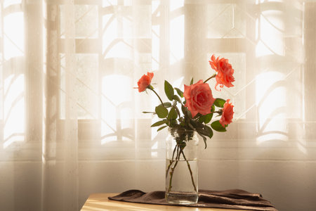 Pink roses in a vase placed on wooden table by window with white curtains, create moody tone and...